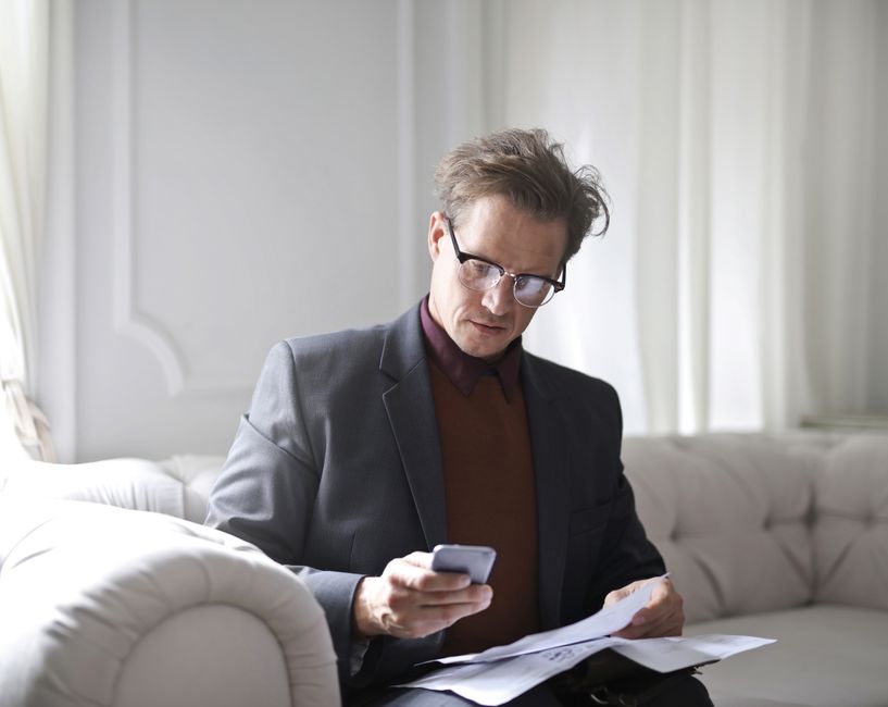 Focused businessman on a sofa using smartphone and reviewing documents.