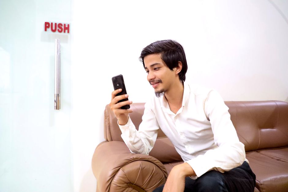Young man in white shirt smiling at smartphone while sitting on a sofa indoors.